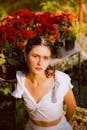 Woman in white dress posing by vibrant red flowers outdoors, basking in natural sunlight.