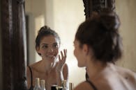 Woman smiling while touching her face in the mirror, enjoying a skincare routine.